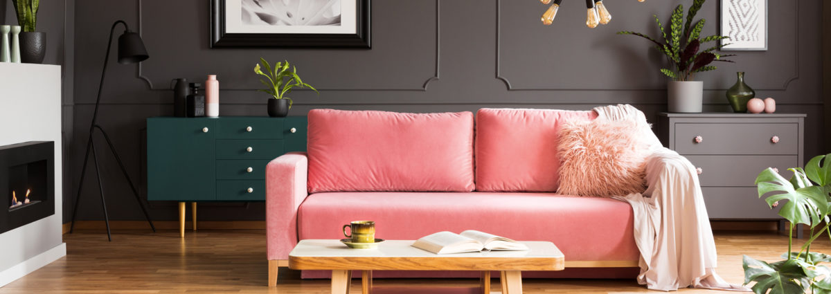 Wooden coffee table with tea cup and open book standing on carpet in dark living room interior with powder pink lounge with fur cushion and blanket
