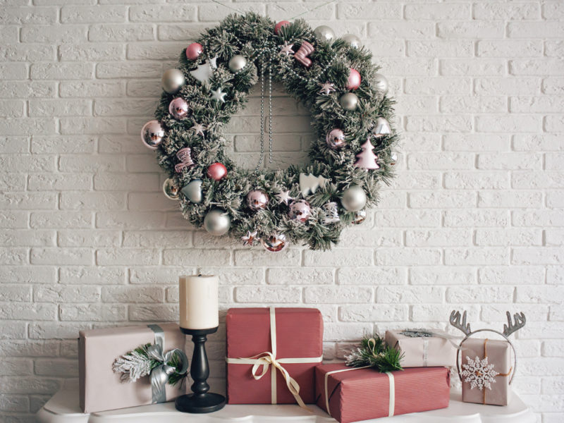 A traditional bright Christmas wreath hanging over the fireplace, on a white brick wall, and packaged gifts are stacked on a fireplace with candles.