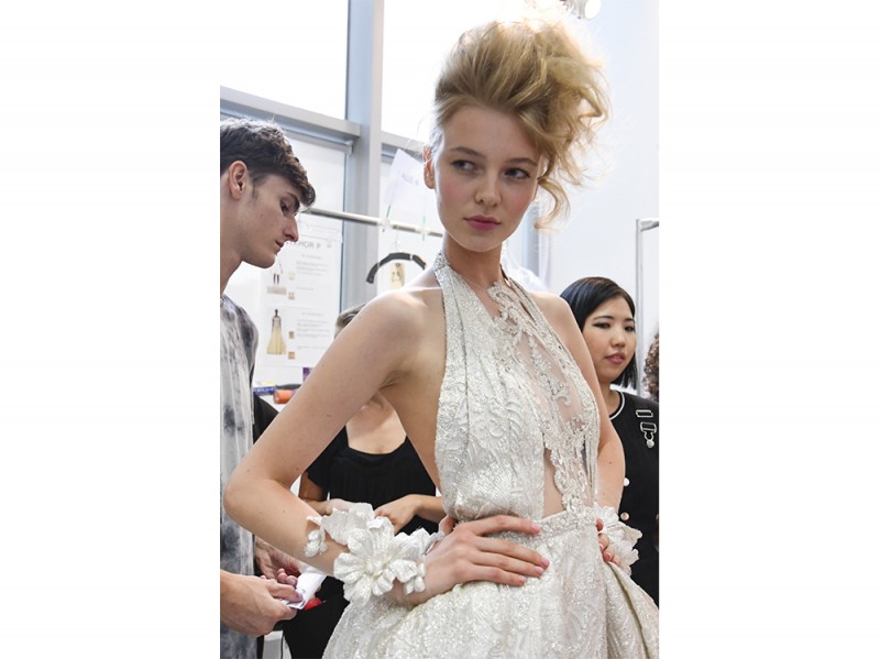 L’acconciatura raccolta si sviluppa in senso verticale e laterale, con onde cotonate che ricadono su un lato del viso.