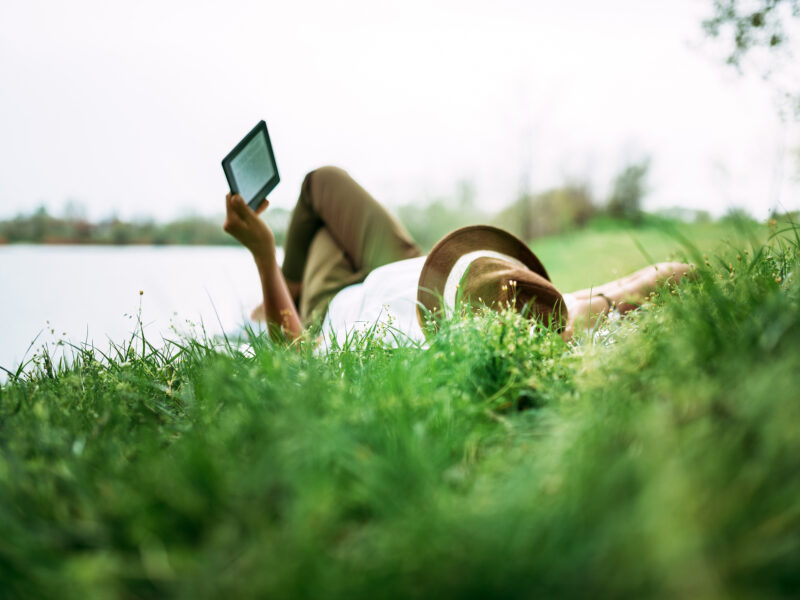 Enjoying e-book near the lake. Girl lying in the grass.