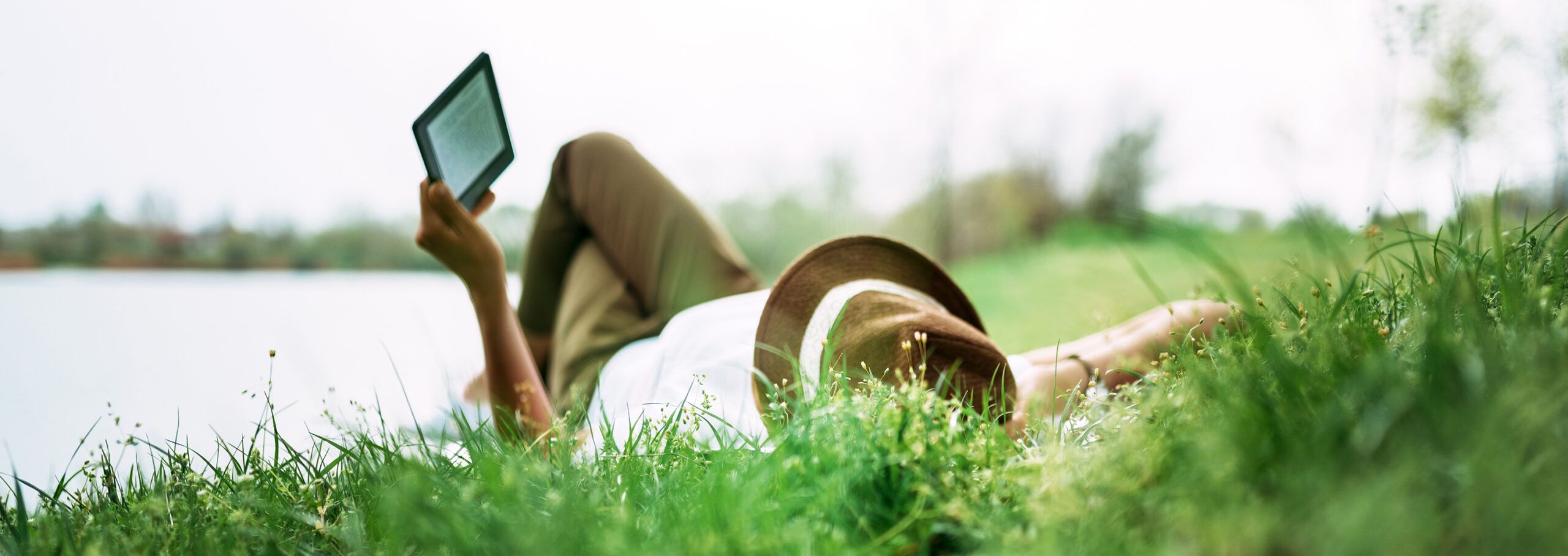 Enjoying e-book near the lake. Girl lying in the grass.