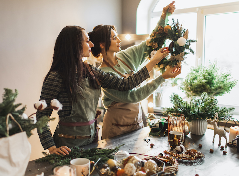 Two women making Christmas wreath using fresh pine branches and festive decorations.