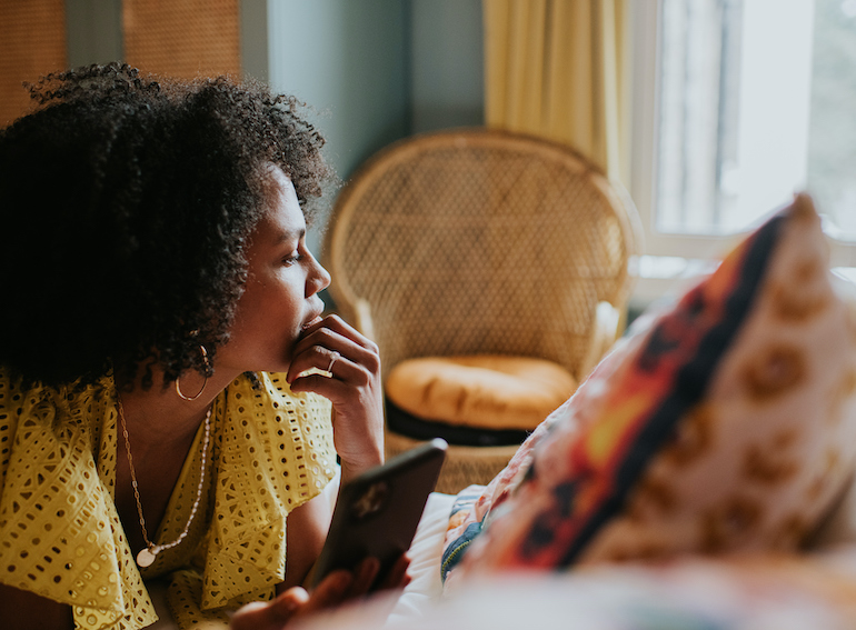 A woman looks distracted and pensive as she lies on a bed holding a mobile phone, and gazes out the window