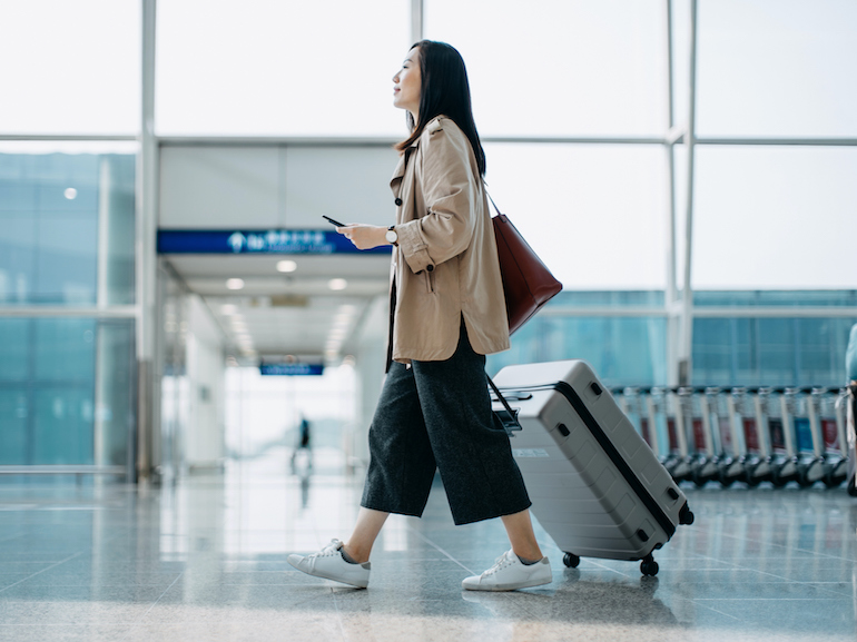 Young Asian woman carrying suitcase and holding smartphone on hand, walking in airport terminal. Ready to travel. Travel and vacation concept. Business person on business trip