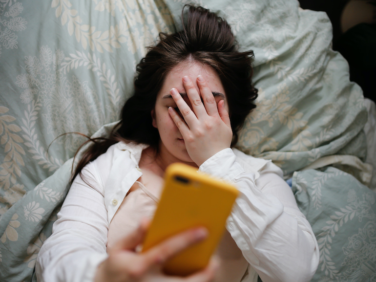 Sleepy overweight young woman with flowing hair using phone on bed, close face palm. Top view