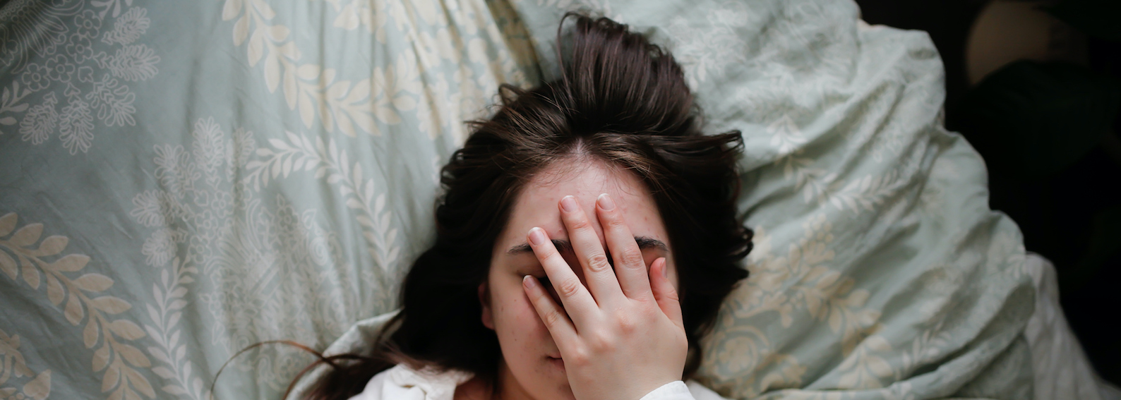 Sleepy overweight young woman with flowing hair using phone on bed, close face palm. Top view