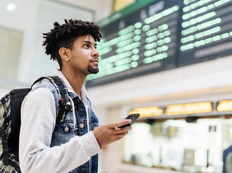 Young man at the airport looking at the list of destinations holding a cell phone.