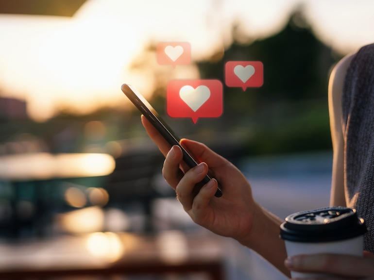 Close up of young woman checking social media network on smartphone on the go in the city. Mobile phone with heart emoji speech bubble message on the screen. People engaging in networking with technology. Social media addiction concept