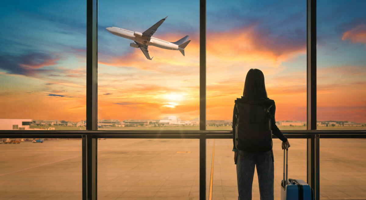 Travel tourist standing with luggage watching sunset at airport window. Woman looking at lounge looking at airplanes while waiting at boarding gate before departure. Travel lifestyle. Transport and travel concept