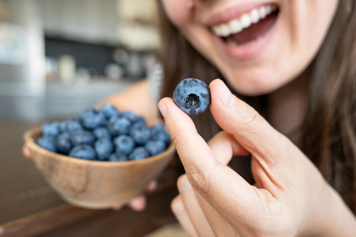 Smiling woman bringing blueberry to mouth with bowl in hand